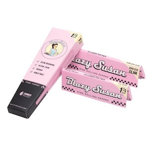 Pode incluir: Papéis de enrolar e cones Blazy Susan cor-de-rosa são exibidos. A embalagem, em rosa com detalhes pretos, apresenta o nome da marca em itálico. O texto inclui "Pink Rolling Papers", "Slow Burning", "Ultra-Thin", "Vegan" e "GMO Free".