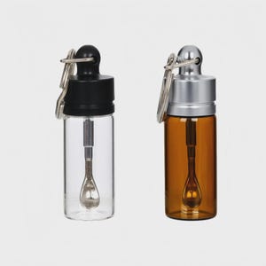 May include: Two small glass vials with attached keychains. One vial is clear with a black cap, the other is amber with a silver cap. Each vial contains a small spoon-shaped tool.