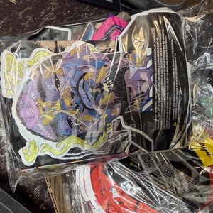 May include: A collection of stickers in clear plastic packaging. The stickers feature colorful illustrations with purple, blue, yellow, and green accents. Some stickers have a comic book style. A black package with text is also visible.