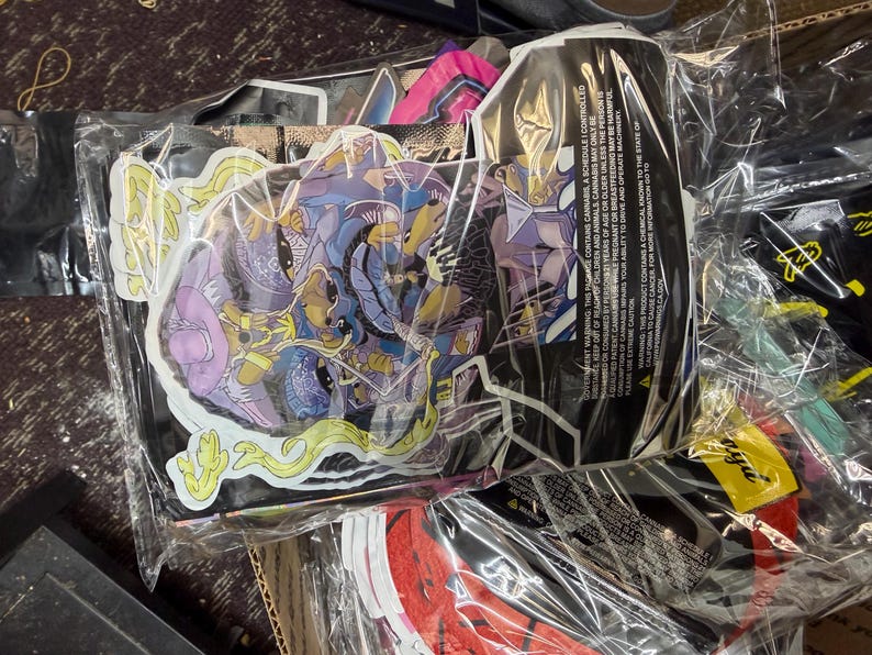 May include: A collection of stickers in clear plastic packaging. The stickers feature colorful illustrations with purple, blue, yellow, and green accents. Some stickers have a comic book style. A black package with text is also visible.