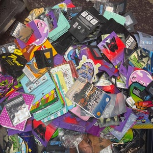May include: A pile of colorful, sealed bags and pouches with various designs and text. The bags are in shades of black, purple, green, and clear, with images and text including "Connected" and "Pigpen". Some bags are sealed with clear plastic.