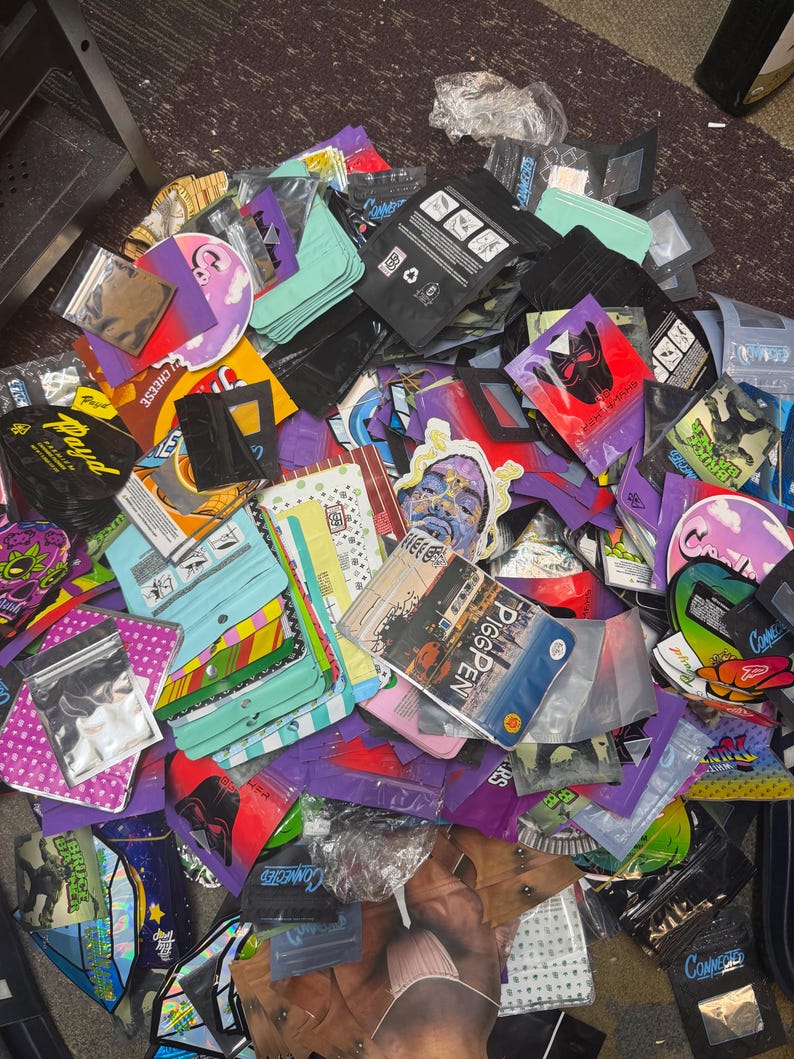 May include: A pile of colorful, sealed bags and pouches with various designs and text. The bags are in shades of black, purple, green, and clear, with images and text including "Connected" and "Pigpen". Some bags are sealed with clear plastic.