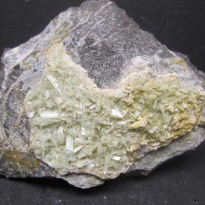 May include: A rough, gray rock with a central cluster of pale green, crystalline formations. The crystals are elongated and clustered, set against a backdrop of lighter, yellowish-brown material. The rock's surface is textured.