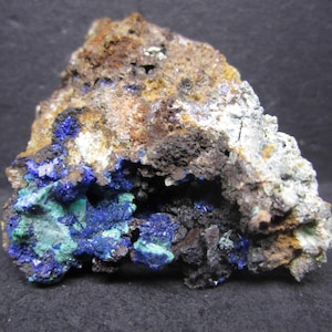 May include: A rough, natural mineral specimen featuring a mix of colors. The rock has a dark brown and tan exterior with patches of blue and green crystals in a cavity. The overall texture is coarse and uneven.