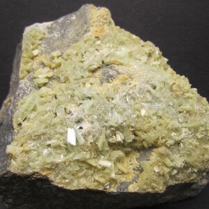 May include: A rough, gray rock formation with a surface covered in clusters of pale green, crystalline structures. The crystals have a slightly translucent appearance, and the overall texture is rough and uneven. The rock is a natural mineral specimen.