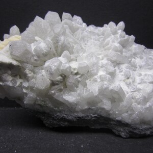 May include: A cluster of white quartz crystals, with a crystalline structure, sits on a dark gray base. The quartz has a translucent appearance, with sharp, pointed terminations. The overall shape is irregular and textured, showcasing natural formations.