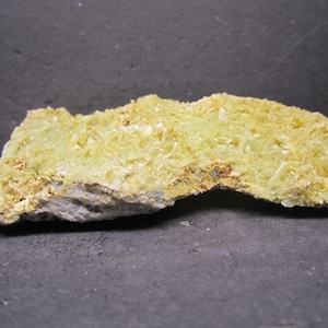 May include: A light yellow mineral specimen with a crystalline structure. The mineral has a rough, uneven surface and is set against a dark gray background. The crystals are small and densely packed, creating a textured appearance.