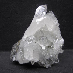 May include: A cluster of clear quartz crystals with a prominent, sharply pointed crystal at the top. The crystals are translucent and sit on a dark, gray-black matrix. The overall shape is irregular and jagged, showcasing natural mineral formations.