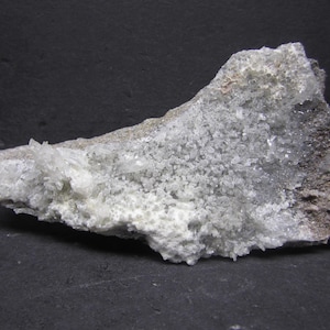 May include: A natural mineral specimen featuring a cluster of small, clear quartz crystals. The rock has a light gray base with a coating of tiny, sparkling crystals. The overall appearance is rough and textured, with a mix of white and gray hues.