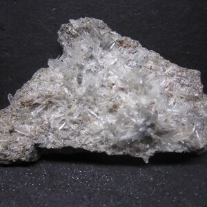 May include: A close-up of a mineral specimen featuring a cluster of clear, needle-like crystals. The crystals are densely packed and protrude from a rough, gray matrix. The background is a dark, textured surface.