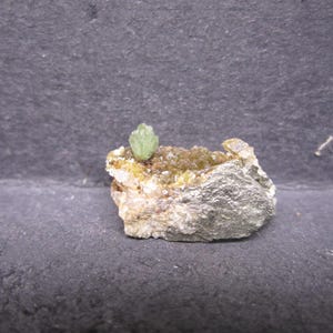 May include: A small, rough rock formation featuring a cluster of small, light green crystals atop a bed of yellow-brown crystals. The rock has a silver-gray metallic appearance.