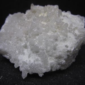 May include: A close-up of a white, crystalline mineral specimen. The mineral has a rough, textured surface with small, pointed crystal formations. The overall shape is irregular, with a slightly rounded appearance. The background is black.