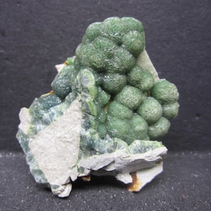 May include: A close-up of a green mineral specimen. The mineral has a bubbly, textured appearance with a light green hue. It sits on a light gray rock base, showcasing its natural form and color variations.