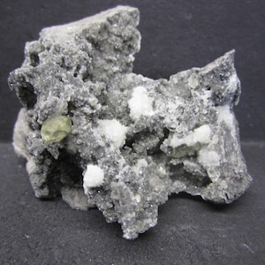 May include: A close-up of a mineral specimen featuring a rough, gray matrix with embedded white and pale green crystals. The texture is granular and uneven, with a dark background.