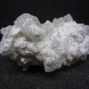 May include: A cluster of light gray quartz crystals. The crystals have a rough, textured surface and are clustered together in a rounded shape. The quartz is set against a dark background.