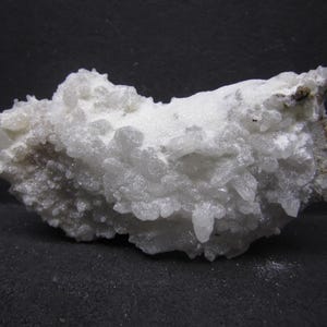 May include: A white, crystalline mineral specimen with a rough, textured surface. The mineral has a cluster of small, pointed crystals and a matte finish. The background is a dark gray.