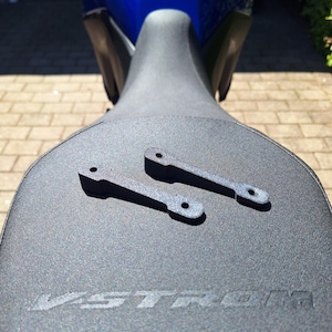 Puede incluir: Dos soportes de motocicleta de metal negro con extremos redondeados y agujeros, colocados sobre un asiento de motocicleta negro. La palabra "V-STROM" está grabada en el asiento. Una motocicleta azul se ve en el fondo.