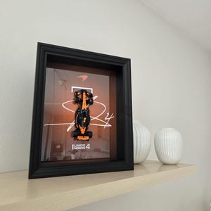 Puede incluir: Obra de arte enmarcada con un coche de carreras de Fórmula 1 negro y naranja, con el nombre "Lando Norris 4". La obra está sobre un fondo naranja y enmarcada en negro.