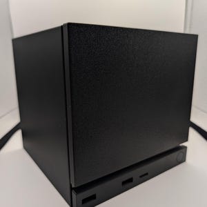 以下が含まれることがあります： 黒色の長方形の電子機器で、表面はテクスチャ加工されています。洗練されたモダンなデザインで、下部には長方形の切り欠きと丸いボタンがいくつかあります。全体的な美しさはミニマリストでインダストリアルです。