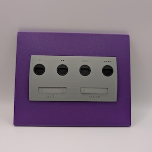 May include: A gray plastic GameCube controller port replica mounted on a purple square. The replica has four circular ports and two slots labeled "SLOT-A" and "SLOT-B". The purple square has a textured surface.