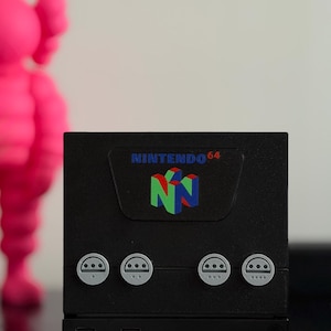 May include: A black, box-shaped video game console featuring the Nintendo 64 logo in red, green, and blue on the front panel. Below the logo are four circular, light gray controller ports arranged horizontally across the front of the device.