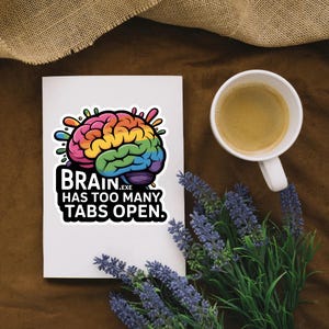 Peut inclure: Un autocollant blanc avec un cerveau coloré et le texte "BRAIN.EXE HAS TOO MANY TABS OPEN." est présenté. Une tasse de café et un bouquet de lavande sont également visibles.