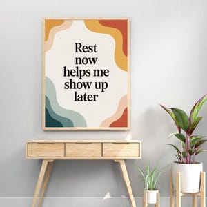 Peut inclure: Une affiche encadrée avec le texte "Rest now helps me show up later". L'œuvre présente un fond crème avec des formes abstraites colorées en orange, rouge, bleu et pêche. Le cadre est de couleur bois clair. L'affiche est exposée sur une table en bois.