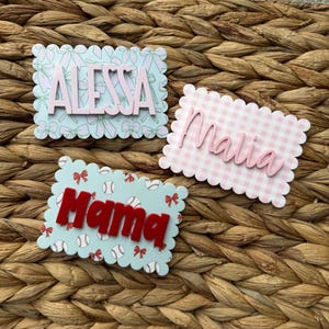 May include: Three rectangular scalloped-edge plaques featuring raised lettering. One plaque has a light blue background with green bows and the name ALESSA in pink. Another has a pink gingham pattern with the name Malia in pink. One has a light blue background with baseballs and red bows with the name Mama in red.