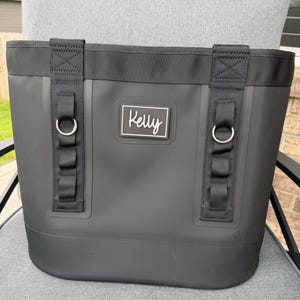 May include: A black tote bag with a rectangular white label that reads "Kelly". The bag has black straps and silver-colored metal rings. The bag is sitting on a gray chair.