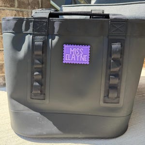 May include: A matte black tote bag featuring two sturdy black nylon handles and vertical webbing straps on the front. A rectangular purple patch with a scalloped edge is centered on the bag, displaying the text MISS ELAYNE in white letters.