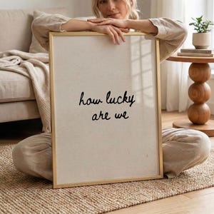 May include: A framed art print with the text "how lucky are we" in black script. The frame is gold, and the background is a light beige. The print is held by a person sitting on a woven rug.
