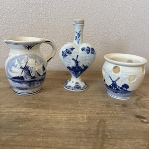 May include: Three Delftware ceramic pieces: a pitcher, a heart-shaped vase, and a small pot. Each piece is white with blue windmill and landscape designs. The pitcher has a handle, the vase has a pedestal base, and the pot has circular cutouts.