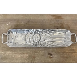May include: A rectangular silver serving tray with handles. The tray features a detailed sun and crescent moon design in the centre. The handles have a textured, rope-like appearance. The tray is on a wooden surface.