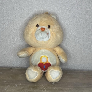 May include: A tan Care Bear plush toy with a heart-shaped nose and a heart-shaped lock and key design on its belly. The plush has a white muzzle and heart-shaped paws. The bear is sitting on a wooden surface against a textured wall.