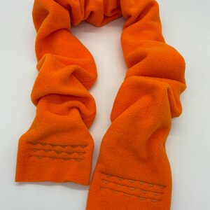 以下が含まれることがあります： 鮮やかなオレンジ色のフリーススカーフ。端には波模様の刺繍が施されています。柔らかく、ふわふわした素材で作られており、首に巻いて着用するようにデザインされています。冬の暖かいアクセサリーです。