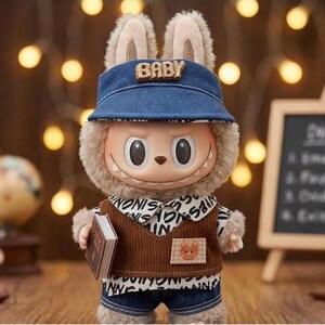 Puede incluir: Una mu&ntilde;eca caprichosa con apariencia de conejo, que lleva un sombrero de mezclilla azul con "BABY", un chaleco marr&oacute;n y pantalones cortos de mezclilla. La mu&ntilde;eca sostiene un peque&ntilde;o libro y tiene una expresi&oacute;n amigable. El atuendo de la mu&ntilde;eca tiene un parche de oso.