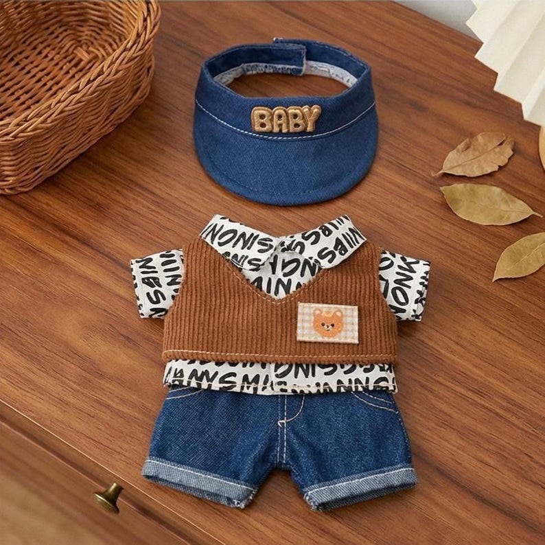 Puede incluir: Un conjunto para beb&eacute; que incluye una visera vaquera con la palabra "BABY" bordada, un chaleco de pana marr&oacute;n y un conjunto de pantalones cortos vaqueros. El chaleco tiene un parche de oso y est&aacute; superpuesto a una camisa con un patr&oacute;n de texto repetido.