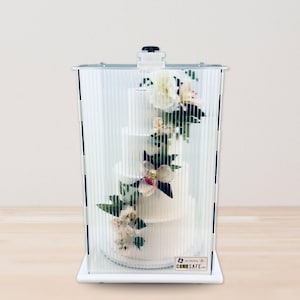May include: A three-tiered white wedding cake decorated with flowers, inside a clear, protective cake carrier. The carrier has vertical lines and a black handle on top. The cake is on a white base.