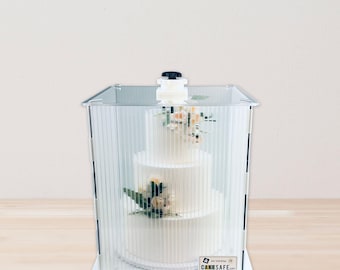 May include: A three-tiered white wedding cake with floral decorations is enclosed in a clear, ridged plastic cake carrier. The carrier has a white base and a black handle on top. The logo for CAKESAFE.com is visible on the front.