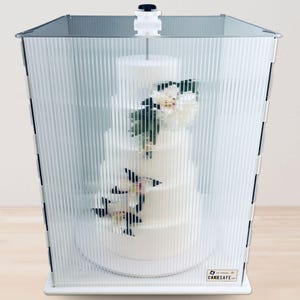 May include: A white tiered cake with floral decorations is encased in a clear, ridged plastic cake protector. The protector has a white base and a handle on top. The cake is on a white surface. The text "CAKESAFE.com" is visible.