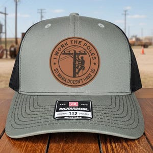 May include: Gray and black trucker hat with the text "I WORK THE POLES SO MAMA DOESN'T HAVE TO" on a brown patch. The hat has a curved brim and an adjustable strap.