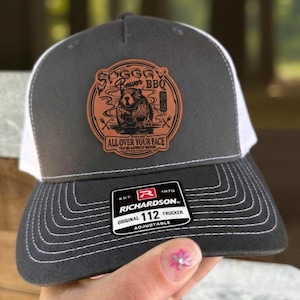 May include: A gray and white trucker hat with a Richardson patch. The hat features a brown leather patch with the text "Soggy Beaver BBQ" and a beaver graphic. The text "ALL OVER YOUR FACE" is also on the patch.