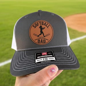 May include: A gray and white trucker hat with a brown leather patch featuring the words "Softball Dad" and a softball player silhouette. The hat has a curved brim and an adjustable strap.