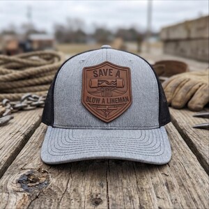 May include: A heather gray baseball cap with black mesh back panels and a brown leather-like shield patch on the front. The patch features an embossed graphic of electrical equipment and the text "SAVE A BLOW A LINEMAN" in capital letters.