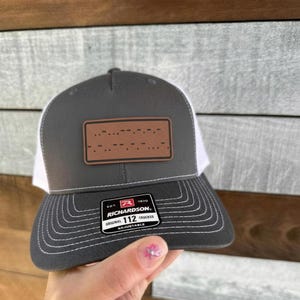 May include: A gray and white trucker hat with a brown rectangular patch featuring a Morse code design. The hat has a curved brim and an adjustable strap. The Richardson 112 hat is a classic style.