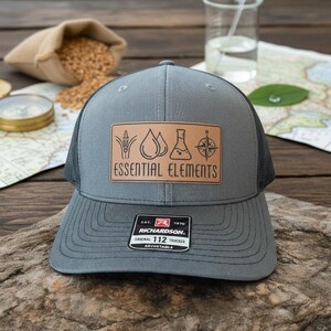 May include: Gray trucker hat with a black mesh back and a brown leather patch. The patch features the words "ESSENTIAL ELEMENTS" and various graphic symbols. The hat has a curved brim and an adjustable strap.
