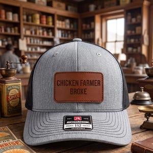 May include: A grey and black trucker hat featuring a rectangular brown leather patch on the front with the text CHICKEN FARMER BROKE engraved in black. The hat includes a black sticker on the brim with the text RICHARDSON EST 1970.
