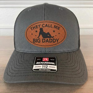 May include: Gray trucker hat with a black mesh back and a curved brim. The front features a brown leather patch with the text "THEY CALL ME BIG DADDY" and a silhouette. The hat is adjustable.