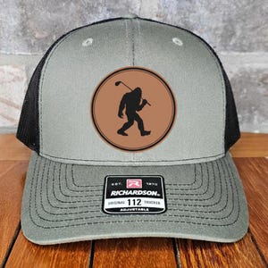 May include: A gray and black trucker hat with a patch featuring a silhouette of Bigfoot playing golf. The patch is brown with a black outline. The hat has a curved brim and an adjustable strap.