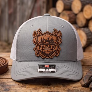 May include: A gray and white trucker hat featuring a brown leather patch on the front. The patch displays a shield design with a deer, mountains, trees, and the text PAPAW. A black and white sticker on the brim reads RICHARDSON 112 TRUCKER.